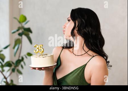 Une femme aux cheveux foncés, portant une robe verte, tient un petit gâteau d'anniversaire avec des bougies '35'. Elle est sur le point de souffler les bougies, un moment joyeux. Banque D'Images