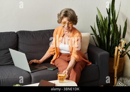 Femme mature est assis confortablement à la maison, sirotant du thé tout en s'engageant avec son ordinateur portable. Banque D'Images