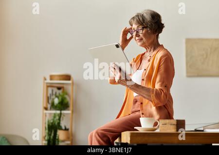 La femme âgée est assise confortablement à la maison, concentrée sur sa tablette avec une tasse chaude à proximité. Banque D'Images