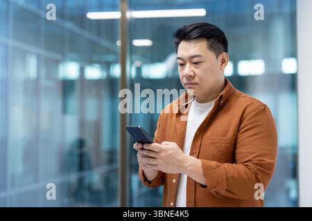 Un homme asiatique utilise son smartphone dans un bureau moderne. Il est habillé de façon décontractée et semble concentré sur son téléphone. Banque D'Images