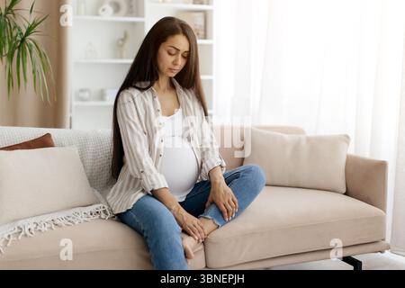 Grossesse problèmes de santé. Jeune femme enceinte en vêtements de maison massant sa cheville gonflée Banque D'Images