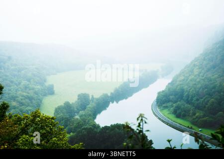 Une vue aérienne d'une rivière sereine serpentant à travers une vallée verdoyante sur un matin brumeux, avec une route paisible le long et des forêts denses. Banque D'Images