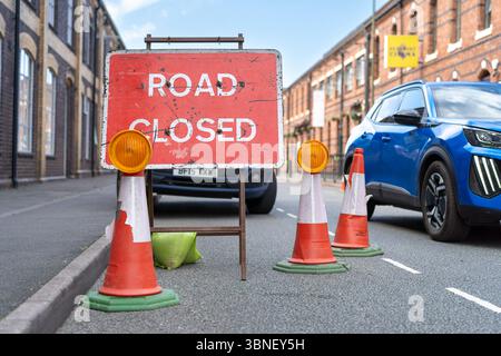 Vue rapprochée et basse d'un panneau rouge « Road Closed » en position sur une route dans le centre-ville du Royaume-Uni près de cônes et de feux d'avertissement pendant que la circulation passe. Banque D'Images