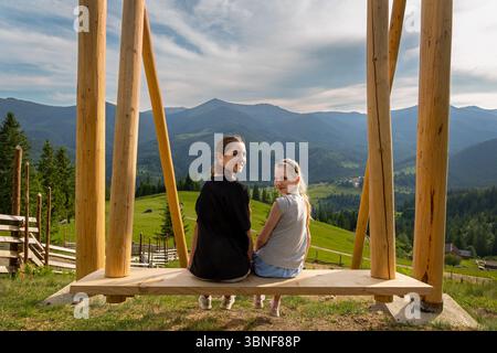 Deux filles assises sur Swing surplombant les montagnes. Deux filles sourient tout en étant assis sur une grande balançoire en bois face à une vue panoramique sur les collines verdoyantes, la forêt Banque D'Images