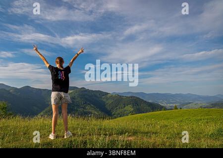 Jeune femme célébrant sur Mountain Viewpoint. Vue arrière d'une femme levant les mains en geste de victoire sur une colline herbeuse, surplombant un vert luxuriant Banque D'Images