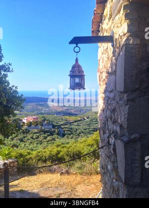 Une lanterne antique est accrochée à un mur de pierre altérée, encadrant une vue tranquille sur la mer et la vallée remplie d'oliviers en Crète, en Grèce. Banque D'Images