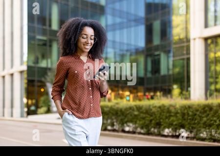 Une femme souriante dans des lunettes utilise son téléphone tout en marchant dehors près d'un immeuble de bureaux moderne. Banque D'Images