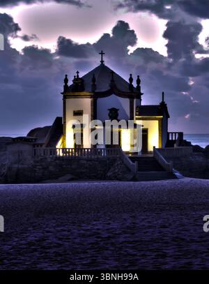 Une église se trouve sur un éperon rocheux au crépuscule, éclairé par des lumières intérieures chaudes. Le ciel est rempli de nuages sombres dramatiques. Banque D'Images