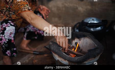 Femme âgée stockant un poêle à bois, préparant un repas traditionnel dans un cadre de cuisine rurale Banque D'Images