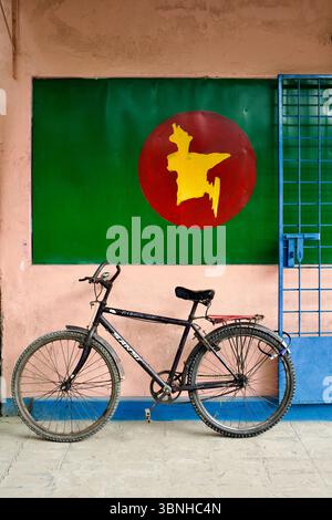 Vélo garé à l'Université de Rangpur devant une murale stylisée du drapeau bangladais avec une silhouette de carte jaune sur un fond vert et rouge. Banque D'Images