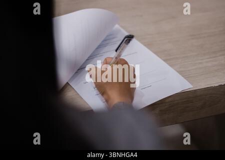 Gros plan d'une main signant un contrat sur un bureau en bois. La personne utilise un stylo pour remplir le document officiel. La prise de vue est nette et précise. Banque D'Images