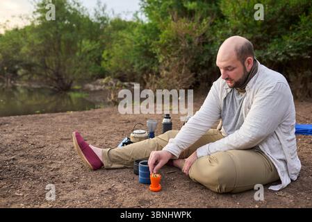 Homme hispanique prenant le petit déjeuner, appréciant une tasse de café fraîchement infusé avec des biscuits sur la rive d'un ruisseau au lever du soleil. Parc national El Palmar, entre Banque D'Images
