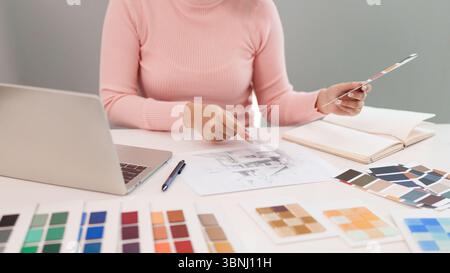 Femme asiatique travaille sur la conception UI UX dans un espace de travail lumineux. Elle sélectionne des couleurs pour un projet de design d'intérieur tout en utilisant un ordinateur portable et une palette de couleurs Banque D'Images