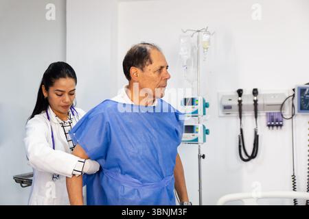 Un médecin assiste un patient portant une robe d'hôpital bleue dans une salle d'examen médical. L'équipement médical et les moniteurs sont visibles en arrière-plan, e. Banque D'Images