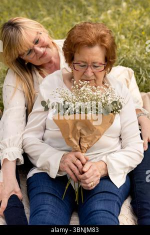 Un moment serein capturant une femme âgée tenant un bouquet tout en étant embrassée avec amour par sa fille adulte. Ils sont assis à l'extérieur, partageant un Banque D'Images