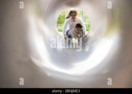Une scène joyeuse capture un bébé garçon et sa mère jouant dans un parc. Ils glissent dans un tunnel de terrain de jeu ensemble, riant et montrant la joie o Banque D'Images