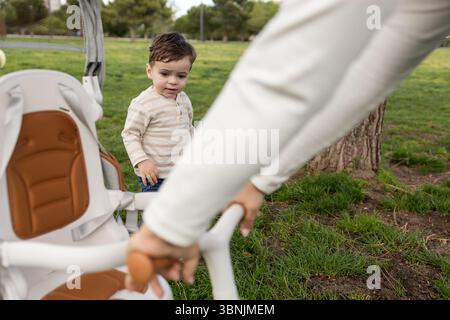 Un tout-petit joyeux se tient dans un parc verdoyant, profitant de temps de qualité avec sa mère. La journée lumineuse et le cadre naturel créent une atmosphère sereine pour Banque D'Images