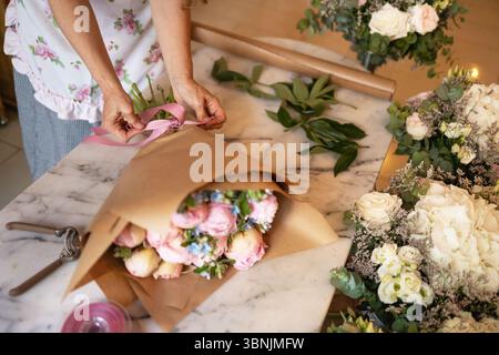 Une femme arrange habilement un bouquet de roses roses sur une table en marbre dans un magasin de fleurs. Elle attache un ruban rose autour de l'arrangement, entouré de b. Banque D'Images