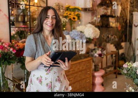 Une femme souriante dans un magasin de fleurs confortable tient une tablette tout en étant entourée de fleurs vibrantes. L'ambiance chaleureuse met en valeur des arrangements florissants et rustiques Banque D'Images