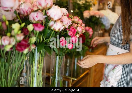 Une femme organise une belle exposition de fleurs dans un charmant magasin de fleurs, organisant des roses roses roses fraîches dans des vases en verre. La scène respire la chaleur et la cre Banque D'Images