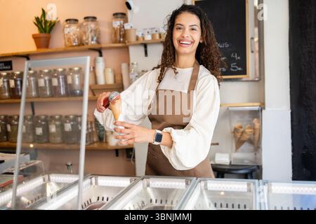 Une femme joyeuse travaillant dans un magasin de crème glacée, servant une boule de délicieuse crème glacée dans un cône. Elle porte un tablier marron et se tient derrière le comptoir Banque D'Images