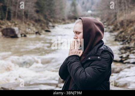 Un homme dans une veste sombre et un sweat à capuche se tient près d'une rivière de montagne qui coule, profondément dans la prière et la réflexion, entouré par la beauté tranquille de la nature, symbolizi Banque D'Images