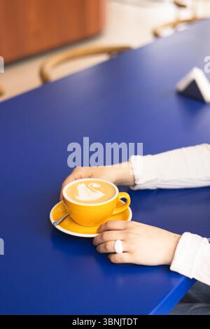 Une femme tient une tasse jaune éclatante ornée d'un dessin de latte complexe en forme de cygne, posée sur une table de café bleu vif, mettant en valeur l'élégance de l'artisanat du café Banque D'Images