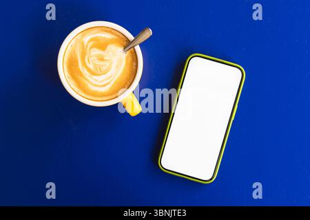 Une image vibrante montre un café fumant dans une tasse jaune à côté d'un smartphone avec un écran vierge, le tout placé contre un plateau bleu saisissant, créant Banque D'Images