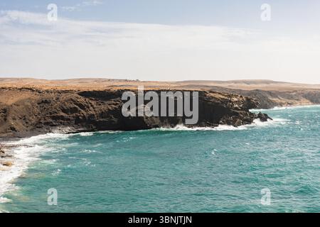 Vue panoramique sur la côte accidentée de Fuerteventura, Espagne, avec des vagues s'écrasant contre les falaises rocheuses sous un ciel bleu clair, offrant un calme Banque D'Images