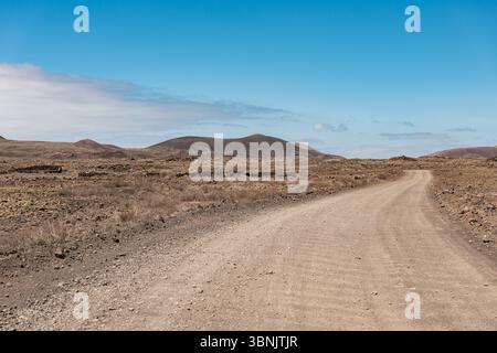 Un chemin de terre sinueux s'étend à travers le paysage aride de Fuerteventura, en Espagne, sous un ciel bleu clair. Collines lointaines et végétation clairsemée complète Banque D'Images