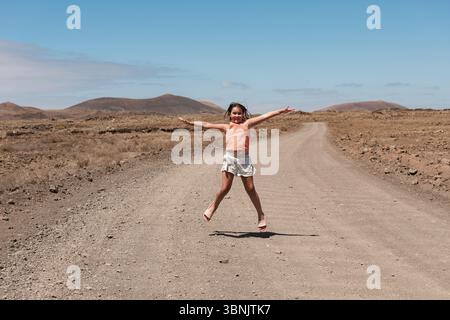 Un enfant joyeux saute les bras grands ouverts sur un chemin de terre déserte à Fuerteventura, en Espagne, entouré de paysages arides et rocheux et de montagnes lointaines U. Banque D'Images