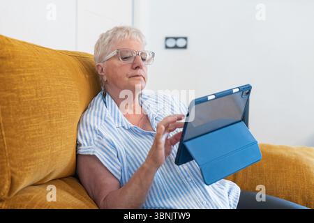 Une femme âgée se détend sur un canapé à la maison, à l'aide d'une tablette. La scène capture l'intersection de l'âge et de la technologie, mettant en évidence l'engagement numérique Banque D'Images