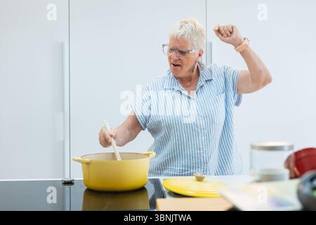 Femme âgée joyeuse avec les bras tendus, cuisinant joyeusement à la maison. Elle exprime le bonheur et l'énergie, capturant l'essence de la cuisine joyeuse M. Banque D'Images