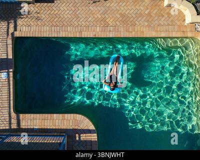 Vue de dessus d'une femme se relaxant sur un radeau gonflable dans une piscine ensoleillée, entourée d'eau claire et pétillante, offrant une ambiance estivale sereine. Banque D'Images