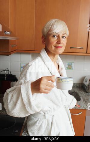Une femme mature aux cheveux courts se tient dans une cuisine moderne, portant un peignoir blanc et tenant une tasse de café. Elle semble détendue et la commence Banque D'Images