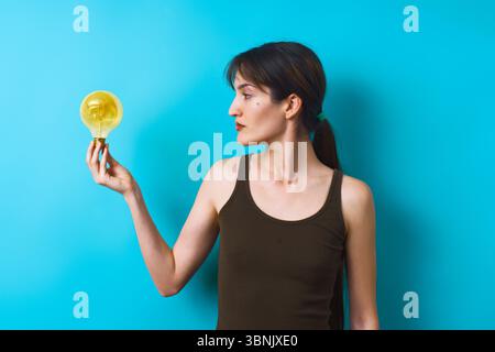 Une jeune femme dans un débardeur sombre tient une ampoule jaune, regardant soigneusement sur un fond bleu vif, symbolisant la créativité et l'innovation Banque D'Images