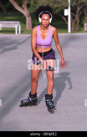 Une sportive mulâtre roller par une journée ensoleillée, portant des écouteurs et des vêtements de sport. Elle tient un smartphone, incarnant un style de vie actif et moderne Banque D'Images
