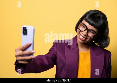 Une jeune femme hispanique dans un blazer violet sourit tout en prenant un selfie avec son smartphone sur un fond jaune vif, exsudant la confiance et Banque D'Images