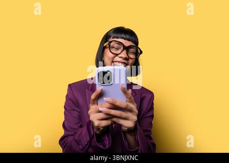 Une jeune femme hispanique joyeuse avec des lunettes sourit largement tout en utilisant son smartphone. Elle est habillée d'un blazer violet élégant contre un cri vibrant Banque D'Images