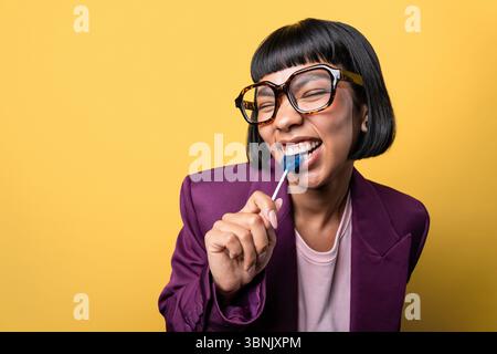 Jeune femme hispanique dans un blazer violet et des lunettes élégantes bénéficie d'une sucette bleue, souriant joyeusement sur un fond jaune vif. La scène Banque D'Images