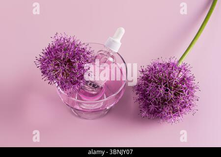 Vue de dessus d'une bouteille transparente de sérum visage placée à l'intérieur d'un bol en verre avec du liquide rose, flanqué de deux fleurs violettes d'allium vibrantes, le tout sur un rose doux Banque D'Images