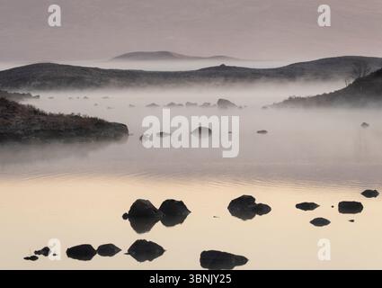 Un paysage serein de Glencoe capturé au lever du soleil, avec un lac couvert de brume parsemé de rochers et entouré de collines doucement vallonnées. Banque D'Images