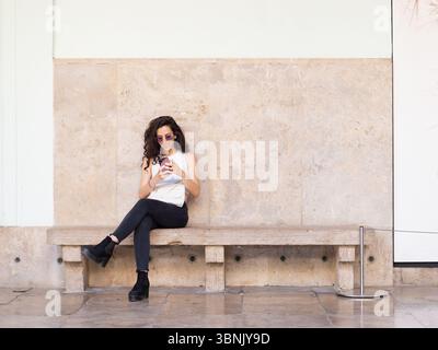 Une jeune femme en lunettes de soleil et en tenue décontractée est assise sur un banc de pierre, absorbée dans son smartphone. La toile de fond urbaine met en évidence un style de vie moderne en t Banque D'Images