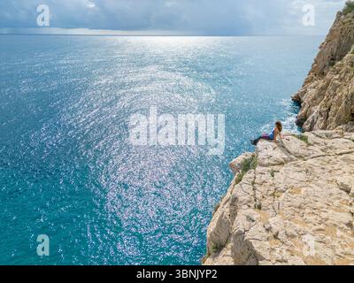 Une jeune femme profite de la vue imprenable depuis une falaise rocheuse sur la Costa Blanca, en Espagne. La vaste mer bleue scintille sous la lumière du soleil, créant un serein Banque D'Images