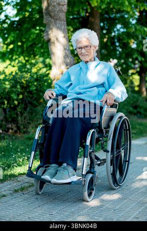 Une femme âgée s'assoit confortablement dans un fauteuil roulant, profitant d'une journée ensoleillée à l'extérieur. Elle est entourée d'arbres verts luxuriants, ce qui crée un calme et s Banque D'Images