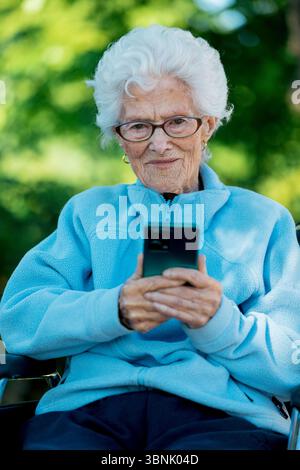 Une femme âgée dans une veste bleue, assise dans un fauteuil roulant, utilise un smartphone avec un sourire paisible. La scène est mise contre un bac extérieur vibrant Banque D'Images