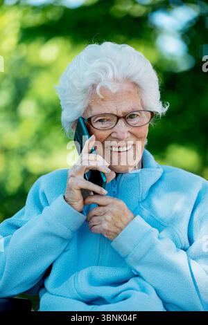 Une femme âgée en fauteuil roulant aime une conversation téléphonique alors qu'elle est assise à l'extérieur. Elle est souriante, portant des lunettes et une veste polaire bleue, avec un Banque D'Images