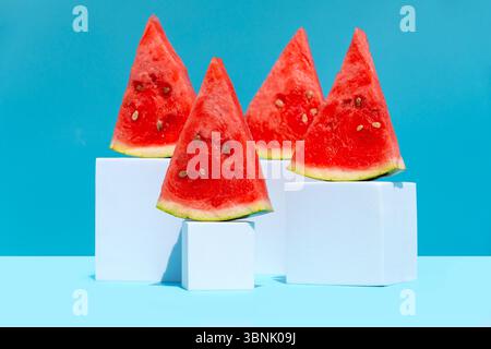 Tranches de pastèque brillantes affichées de manière créative sur des cubes géométriques blancs sur un fond bleu vif. L'arrangement ludique évoque une fraîche et s Banque D'Images