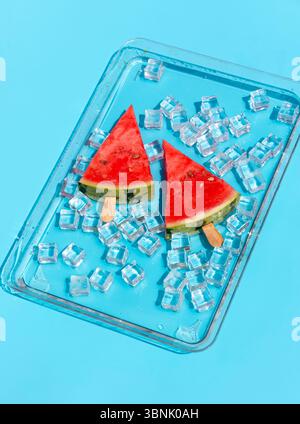 Deux popsicles rafraîchissantes à la pastèque reposent sur un lit de glaçons dans un plateau en verre transparent sur un fond bleu vif, créant un été parfait Banque D'Images