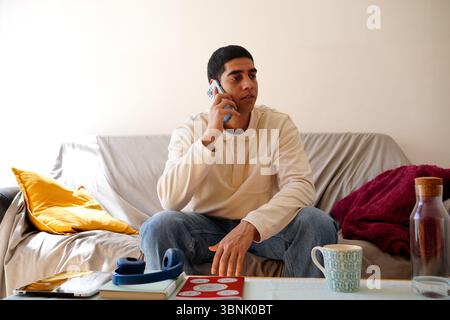 Un jeune homme arabe parlant au téléphone, assis sur un canapé confortable dans sa maison, entouré d'objets du quotidien comme des livres, des écouteurs et une tasse de Banque D'Images